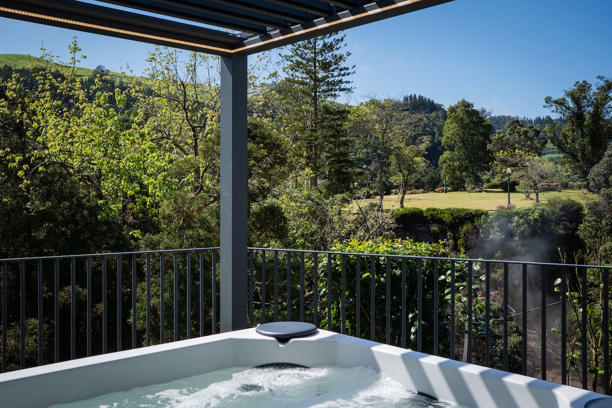 A shared jacuzzi is positioned on a deck with a railing, offering scenic views of lush greenery and hills. Bright blue skies and gentle sunlight enhance the tranquil outdoor setting, creating a serene environment for relaxation.