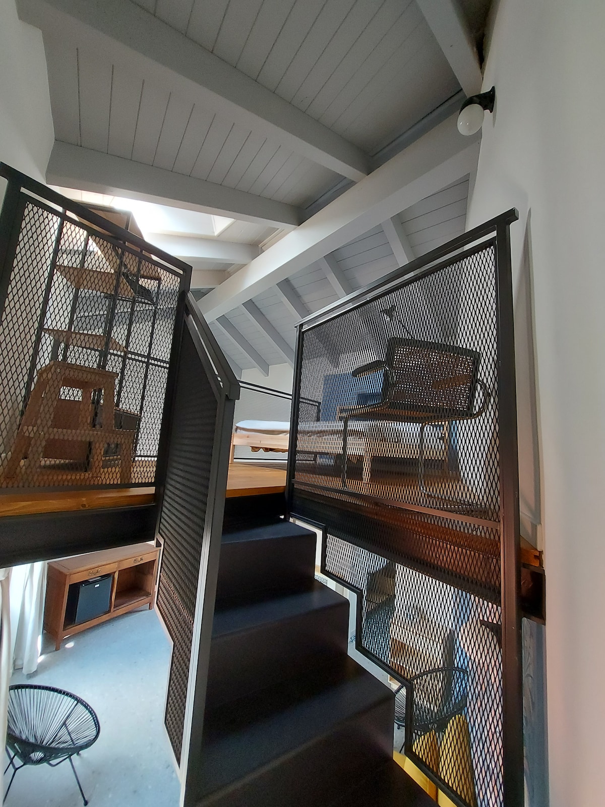 A staircase with metal railings leads to a lofted area, showcasing a spacious design with visible wooden beams overhead. A cozy chair and a bed frame are positioned in the upstairs space, complemented by natural light filtering through a nearby skylight.