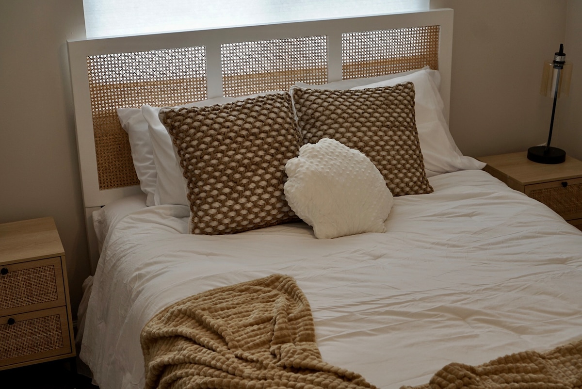 A spacious bed is dressed in crisp white linens, complemented by textured pillows in warm beige tones. A soft throw blanket is casually placed at the foot of the bed. On either side, nightstands add a modern touch, enhancing the serene atmosphere of the sleeping area.
