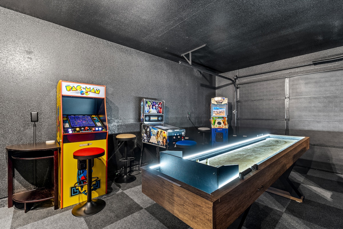 An entertainment room is equipped with an illuminated shuffleboard table at its center, flanked by arcade games such as Pac-Man and a multi-game console. Bar stools surround a small table, offering a casual seating area. The space features dark walls and a high ceiling.