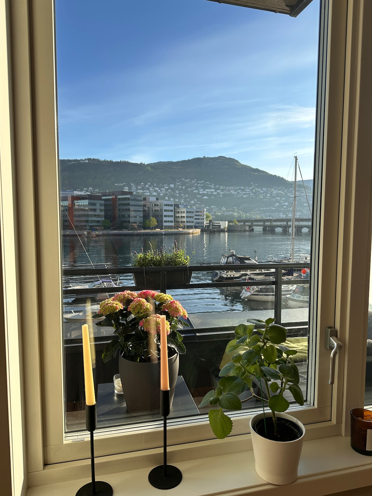 A scenic view of the waterfront is captured through a window, showcasing a peaceful harbor with a mountain backdrop. Potted plants are placed on the windowsill, accentuated by decorative candle holders. The scene reflects a calm atmosphere with boats gently floating nearby.