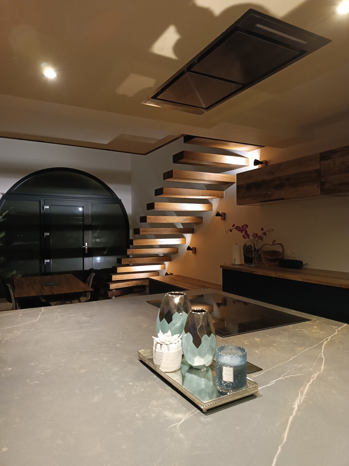 An open kitchen area features a modern design with a large countertop, complemented by wooden shelves and a staircase with wooden treads. Decorative vases and a candle are arranged on a tray, while dim lighting highlights the sleek surfaces.
