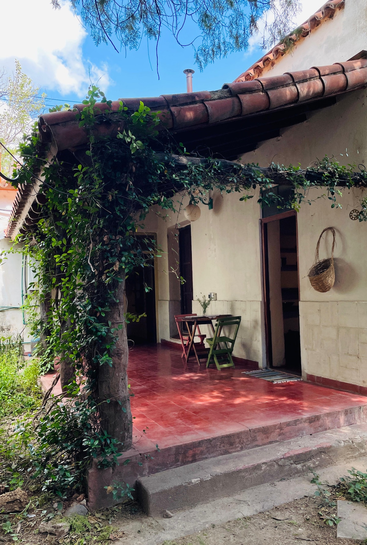 A serene entrance is framed by vibrant greenery, showcasing a welcoming porch with a small wooden table and chairs positioned beneath a sloped, tiled roof. The surrounding area is landscaped with soft earth tones and is illuminated by natural light filtering through the vegetation.