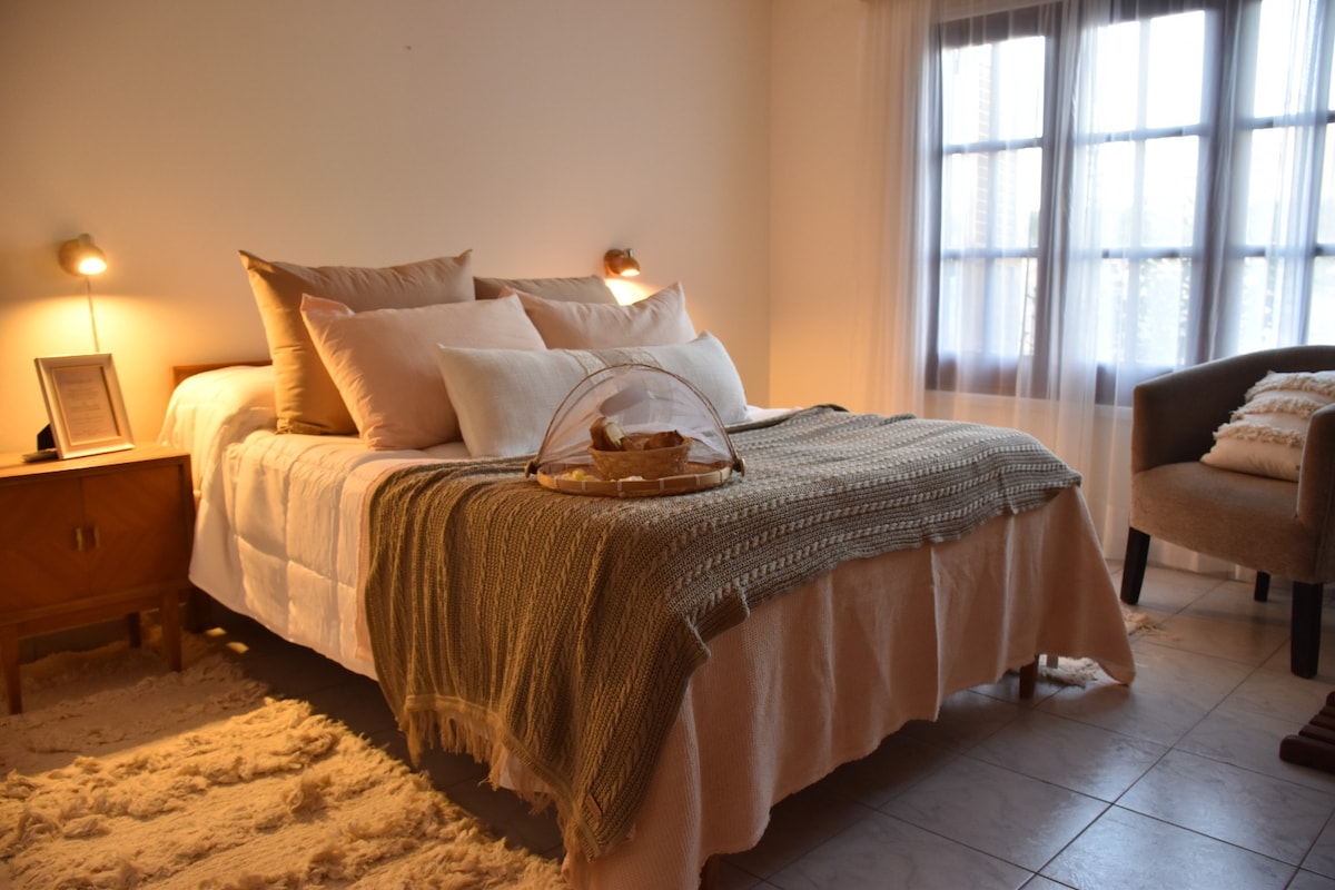 A cozy bedroom is highlighted by a spacious bed with soft, neutral bedding and decorative pillows. A woven throw blanket rests at the foot of the bed. An inviting corner chair and bedside table with ambient lighting complete the tranquil setting, while sheer curtains allow soft light to enter.