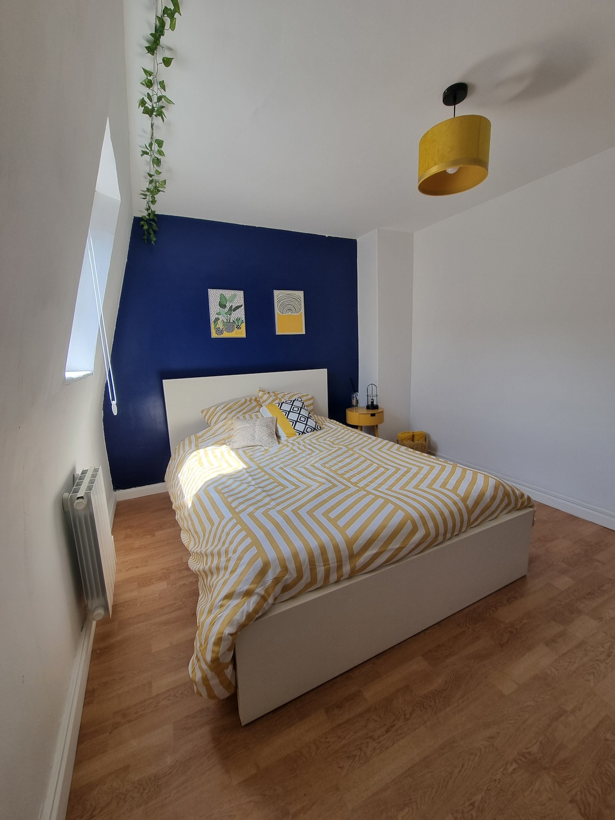 A bright bedroom features a large bed adorned with a yellow and white striped comforter. A blue accent wall enhances the space, while green plants are visible near the window. A matching yellow lampshade complements the decor, creating a cheerful and inviting atmosphere.