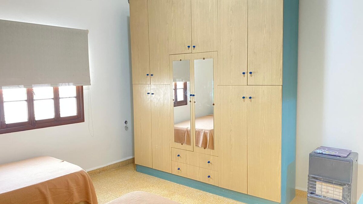 A bright bedroom is depicted, showcasing a light wooden wardrobe with mirrored doors and various storage compartments. A twin bed with a beige bedspread is positioned beside the wardrobe. Soft natural light filters through two small windows adorned with beige shades.