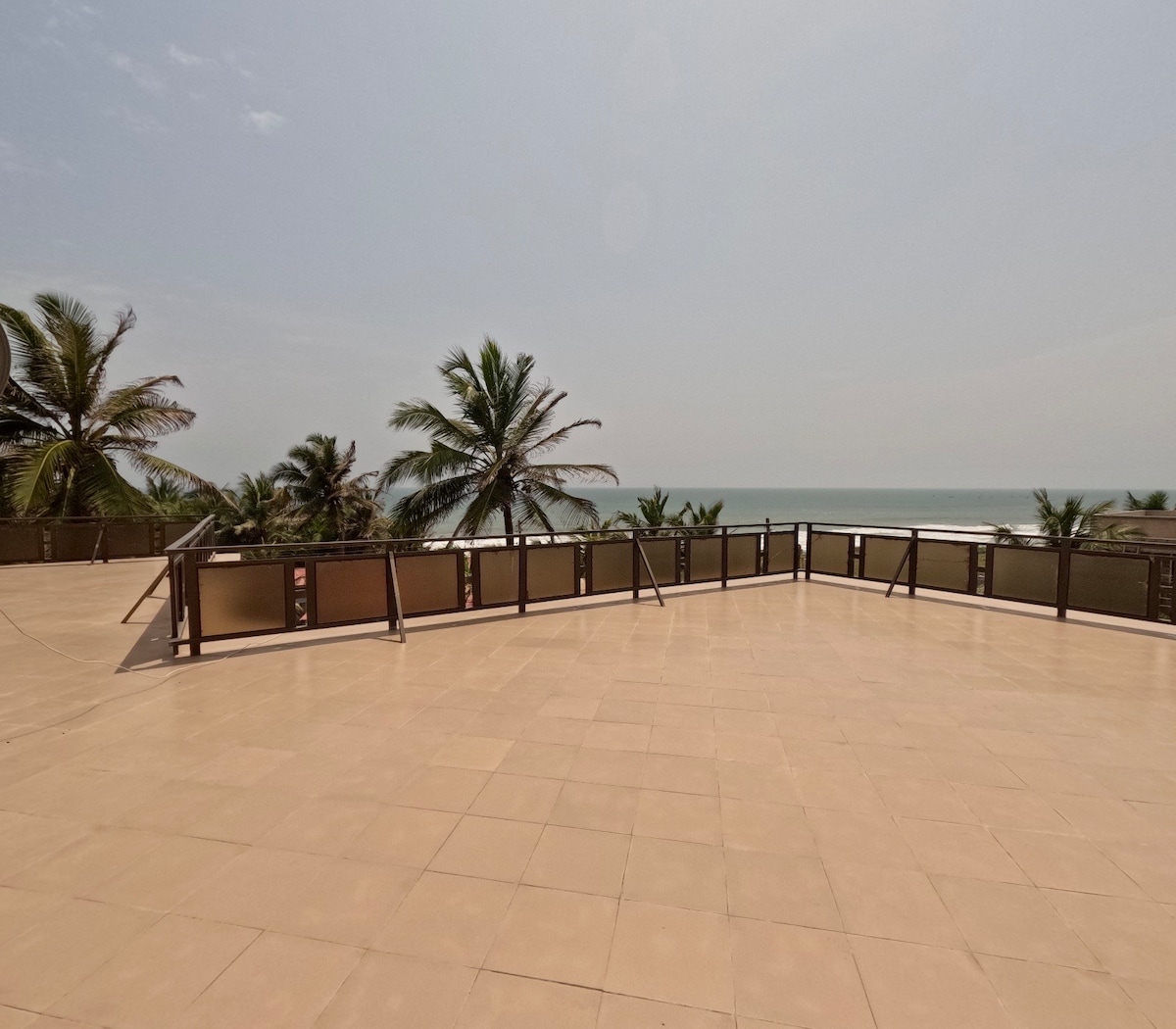 A spacious rooftop terrace features expansive tile flooring and is bordered by sleek railings. Lush palm trees are visible in the background, while the ocean stretches out towards the horizon, creating a serene and open environment for relaxation.