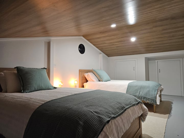 This charming loft bedroom offers a cozy retreat with two comfortable beds, soft linens, and warm wooden accents. The ceiling adds character, while soft lighting creates a relaxing ambiance. Thoughtful storage and plush bedding ensure a restful stay