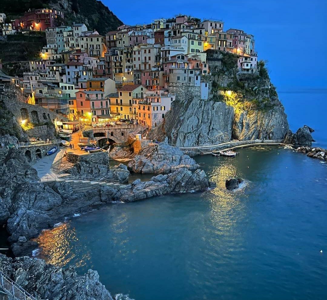 A coastal village is seen illuminated at dusk, with colorful buildings cascading down rocky cliffs. Calm waters reflect the warm glow from street lights and windows. A few boats are anchored near the shore, complemented by rocky outcrops and the distant horizon.
