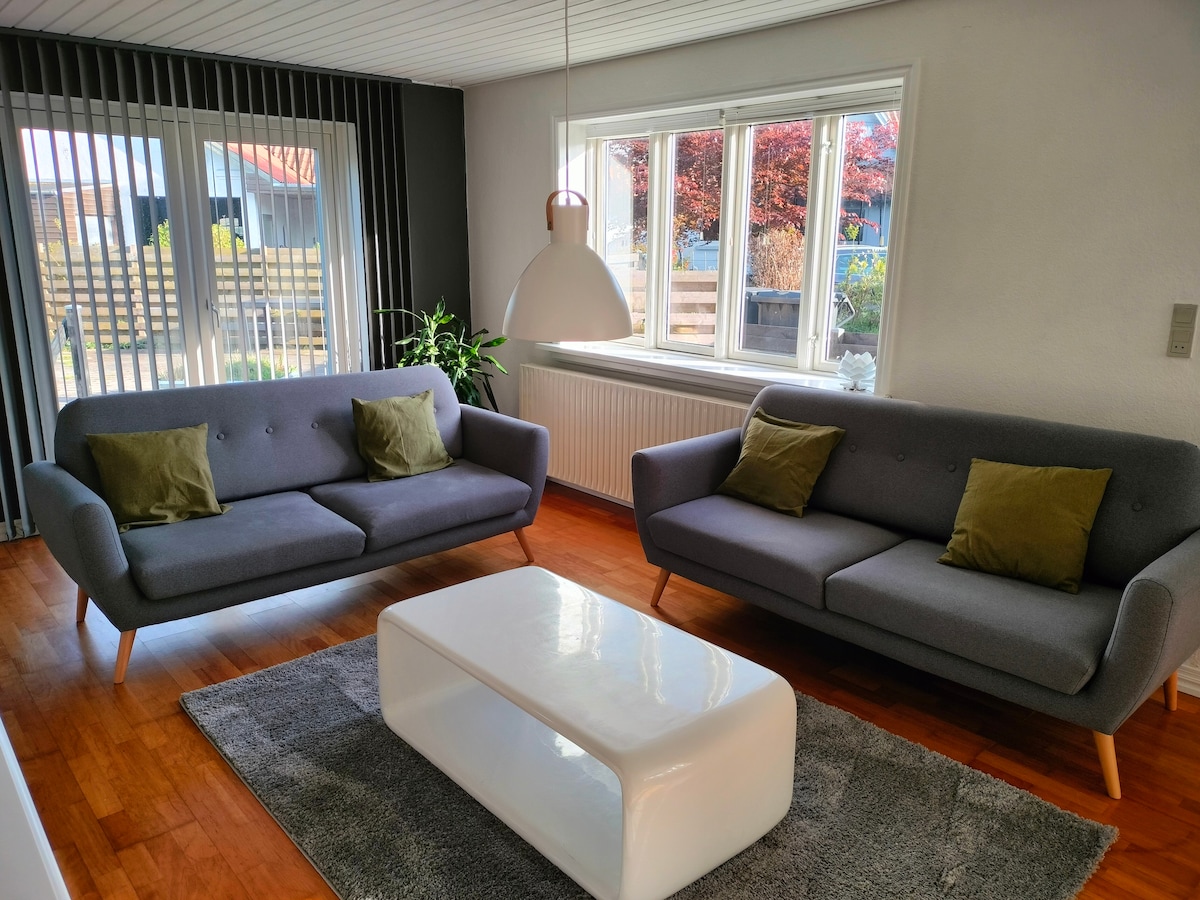 The living area features two modern gray sofas with green cushions, positioned beside a white coffee table atop a light gray rug. Natural light fills the space through large windows, which are adorned with vertical blinds, alongside a decorative plant in the corner.