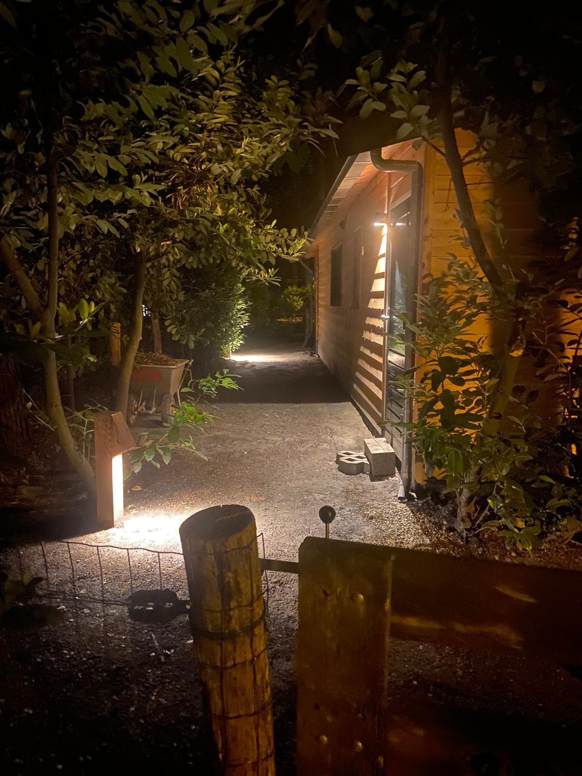A gently illuminated path leads towards the vacation home, surrounded by lush greenery. The warm glow of outdoor lights highlights the natural setting, creating a welcoming entrance. A wooden fence and gate provide access, enhancing the sense of privacy and seclusion.