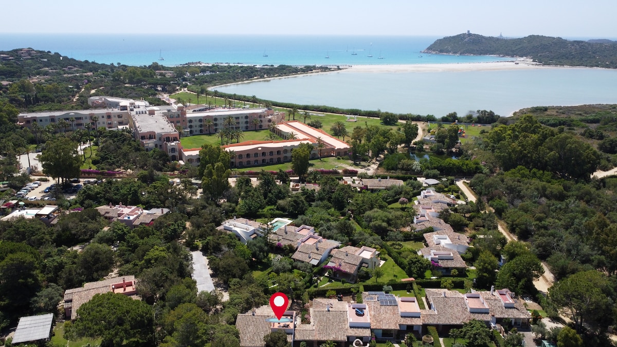 An aerial view captures the villa's surroundings, showcasing lush greenery and sandy dunes alongside a sparkling blue sea. Nearby buildings and the coastline form a picturesque setting, while the location marker highlights the villa's precise position within the tranquil landscape.