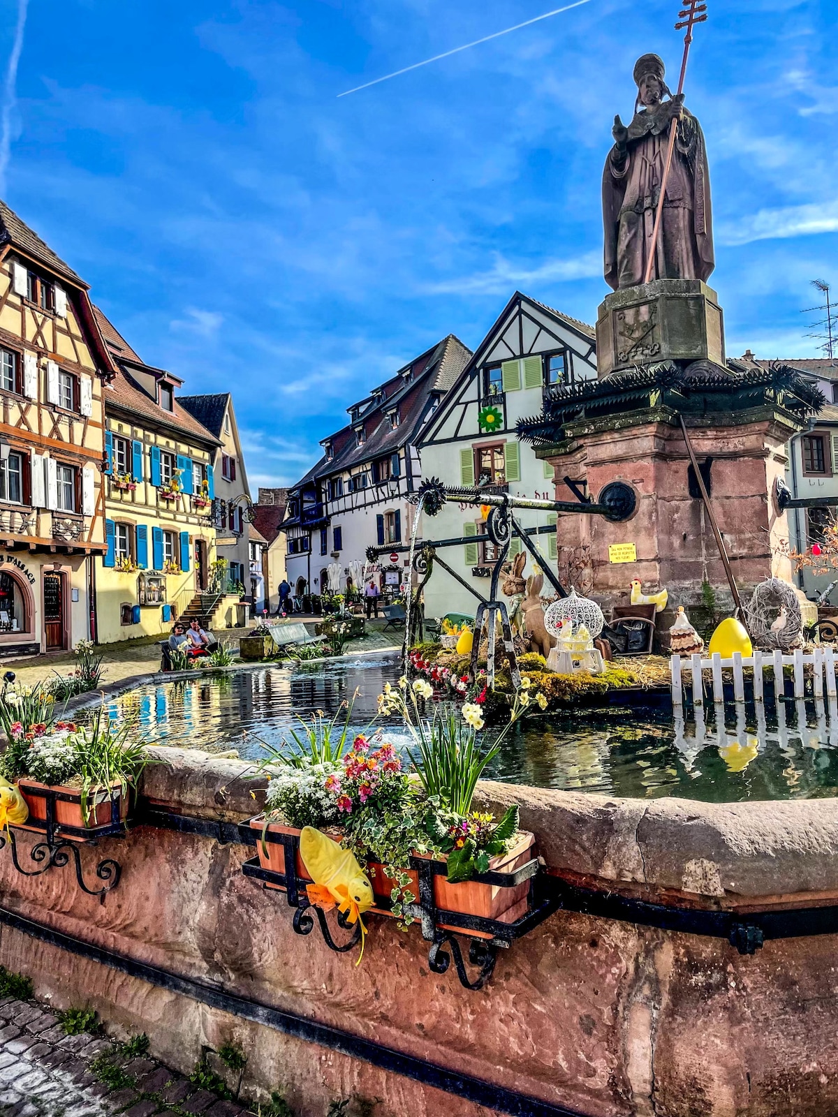 A quaint village square features a decorative fountain surrounded by vibrant flower pots. Colorful half-timbered houses provide a charming backdrop, complemented by a statue on a stone pedestal. The daylight brightens the scene, accentuating the inviting atmosphere of the historic Eguisheim village.