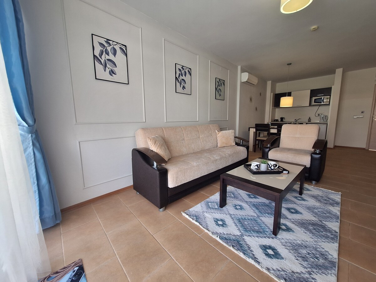 A modern living area is furnished with a light-colored sofa and an armchair, complemented by a coffee table on a patterned rug. Artwork featuring leaves adorns the walls, and natural light filters through sheer curtains, illuminating the space.