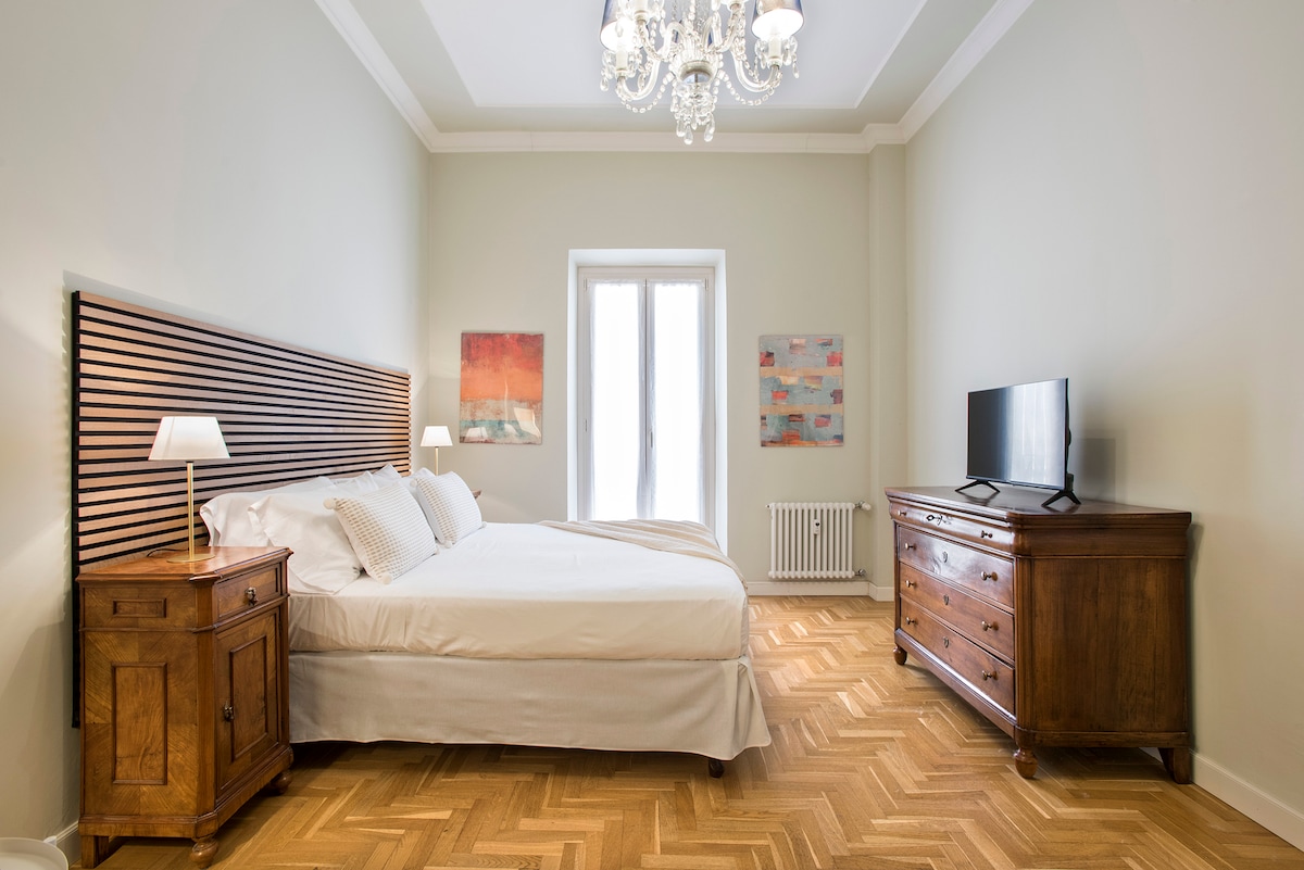 A spacious bedroom features a large bed with a stylish headboard. Two bedside lamps are placed on either side, and a wooden television unit occupies one corner. Natural light floods the room through a large window, complemented by decorative wall art in soft colors.