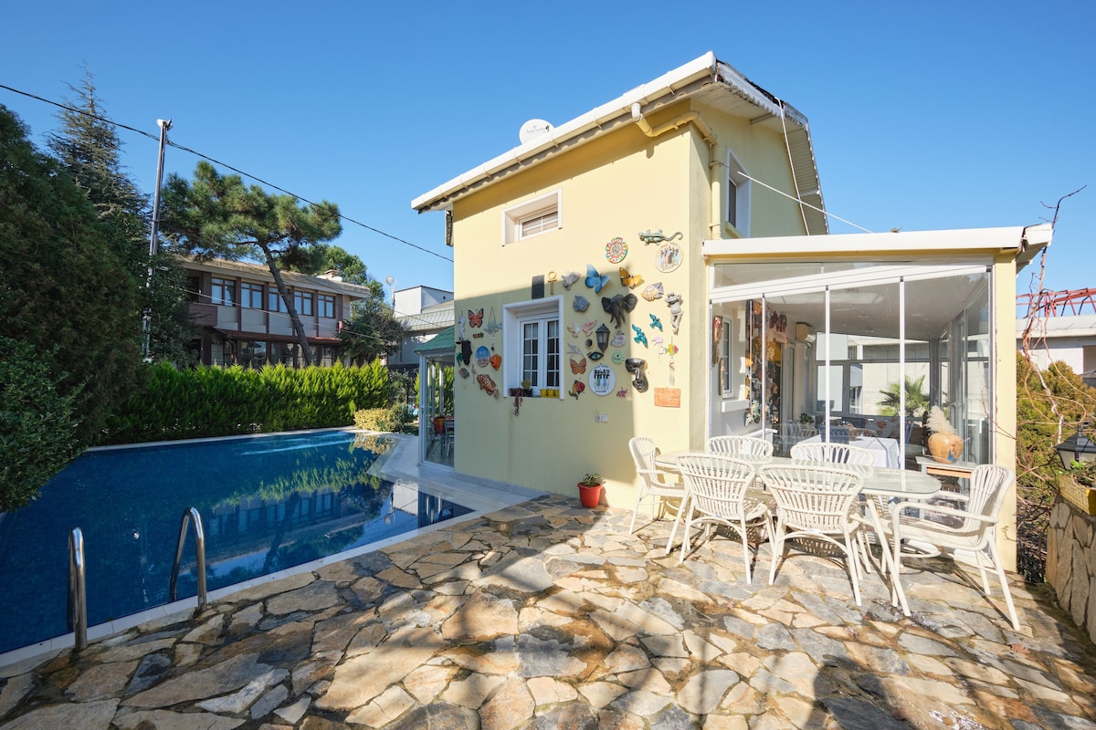 A villa with a light yellow exterior is accompanied by a private pool and a stone patio. White outdoor furniture is arranged for dining beside the water. Colorful wall decorations are visible on the exterior, enhancing the artistic ambiance of the space.
