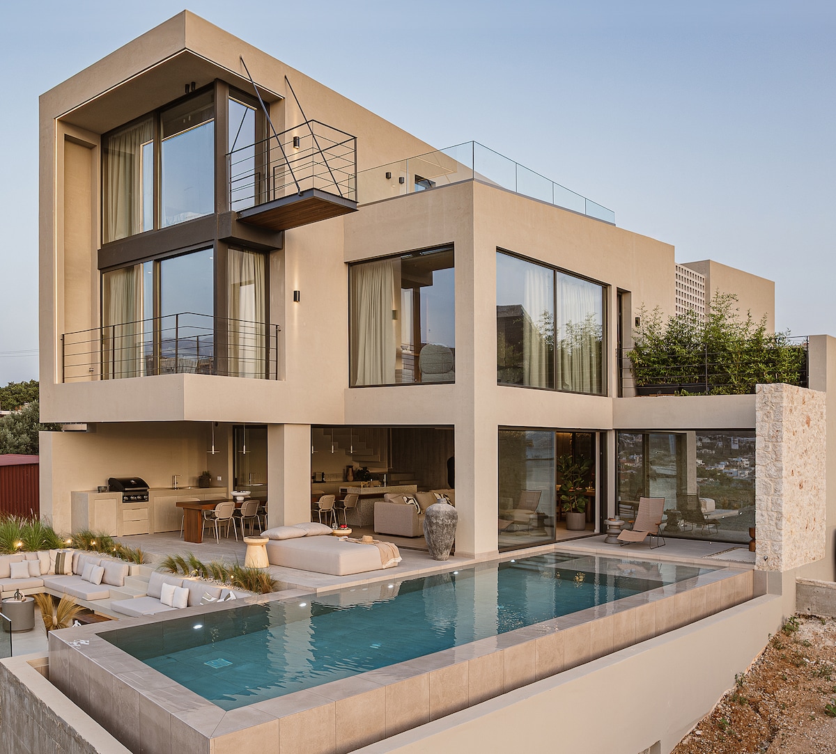 The modern villa features a sleek architectural design with large glass windows and a spacious outdoor area. An infinity pool reflects the sky, while lounge seating is arranged nearby. The minimalist decor suggests a harmonious blend of indoor and outdoor living spaces, inviting relaxation.
