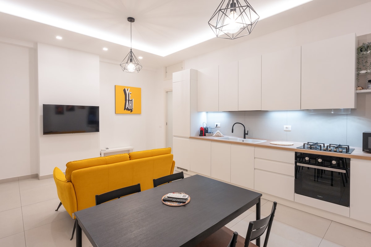 A modern kitchen and living area features sleek white cabinetry and gray countertops. A yellow sofa provides a pop of color, paired with a dark dining table and black chairs. A flat-screen TV is mounted on the wall, illuminated by overhead lighting.