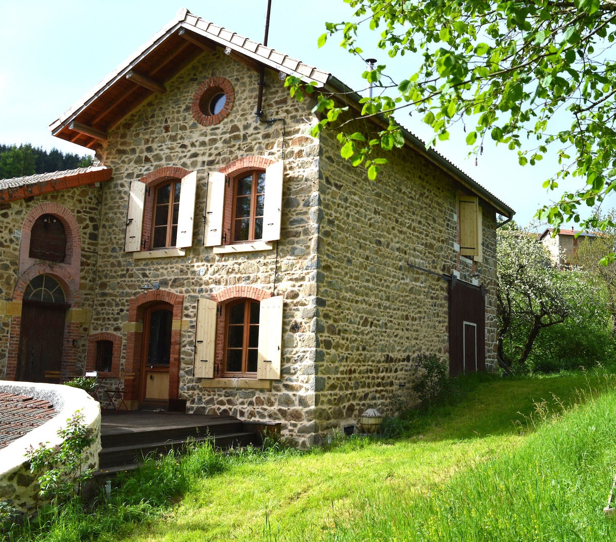 The exterior of a charming stone house is presented, showcasing rustic architecture with wooden shutters and a circular window. A grassy area surrounds the building, complemented by trees and shrubs, highlighting the peaceful countryside setting.