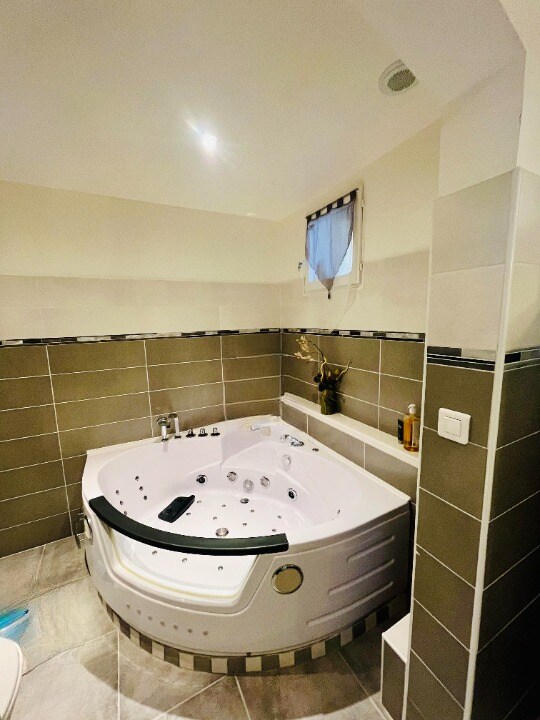 A spacious bathroom features a large whirlpool bathtub, surrounded by modern tile work in neutral tones. A small window allows natural light, and various bath necessities are neatly arranged nearby.