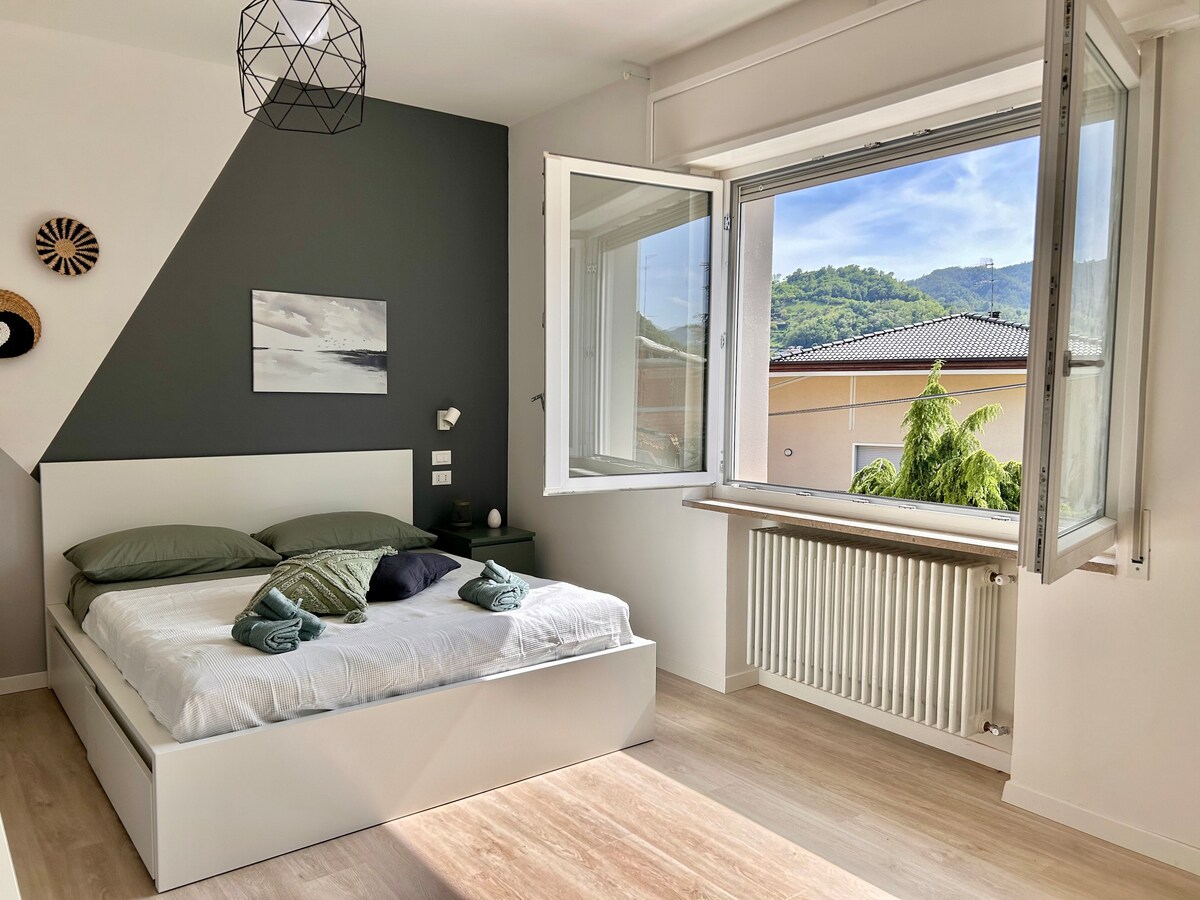 A well-lit bedroom features a spacious bed with soft green linens, complemented by decorative pillows. Large windows allow natural light and fresh air, revealing views of the surrounding greenery and mountains. Minimalist decor and a geometric accent wall add a modern touch to the space.