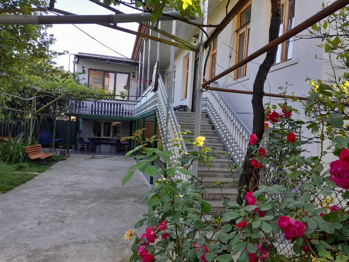 A charming exterior view of the two-story house is presented, featuring a well-maintained garden with vibrant roses. A staircase leads to the second floor, flanked by green foliage and outdoor seating areas below, creating inviting spaces for relaxation.