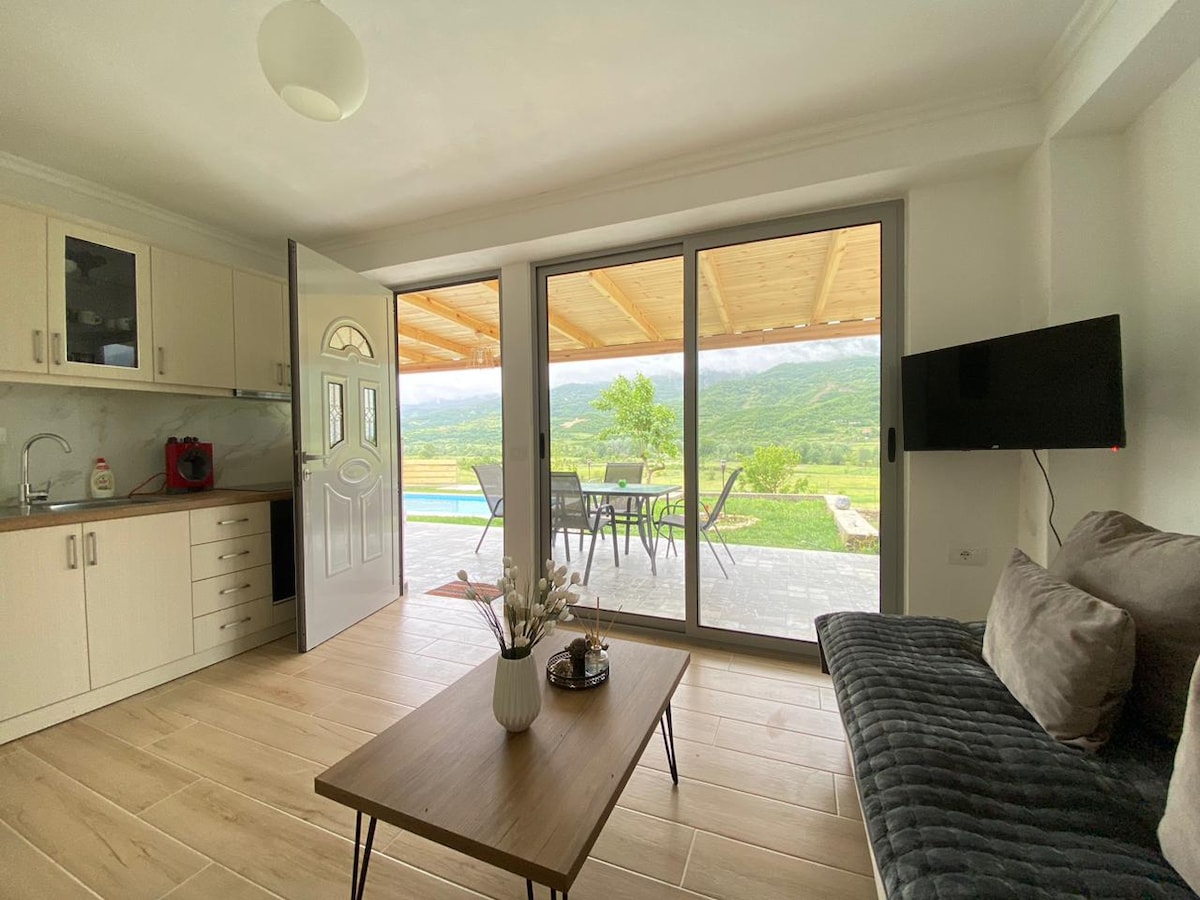 A bright living area features large sliding glass doors that open to a welcoming patio with scenic views of the mountains and valley. A modern kitchenette occupies one side, while a comfortable seating area faces the television, enhancing the space's functionality.
