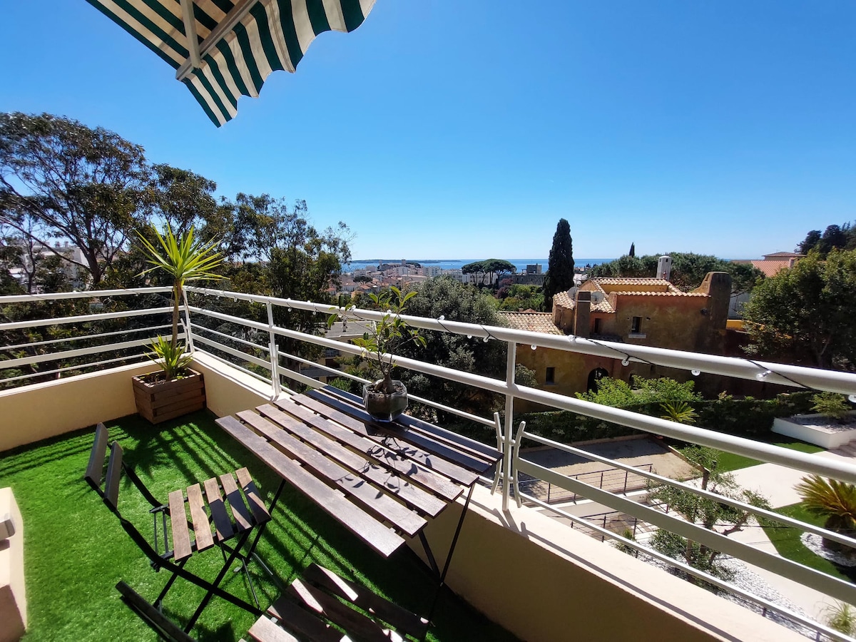 A balcony area features a wooden table and folding chairs, surrounded by potted plants. The view overlooks a scenic landscape with trees and rooftops, leading toward a distant sea. A striped awning provides some shade against a clear blue sky.