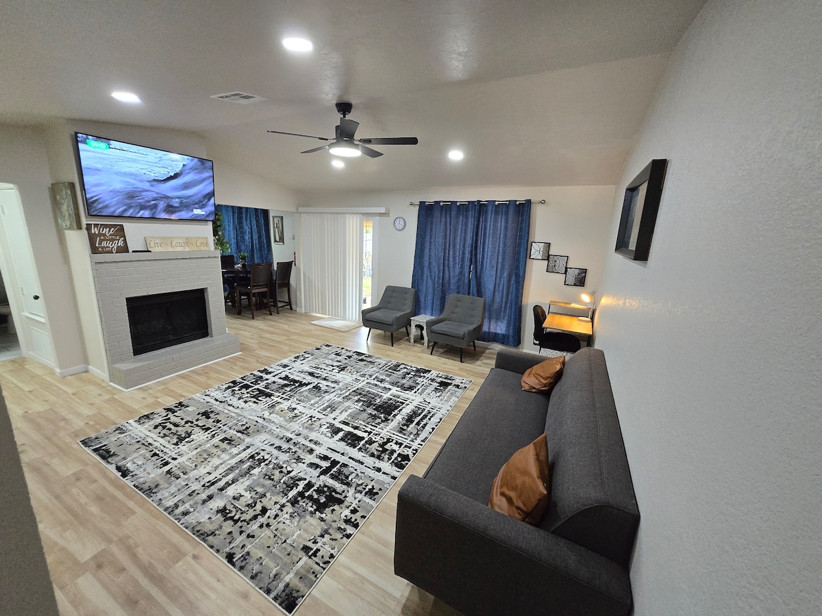 The living area features a dark gray sofa with orange cushions, complemented by two armchairs. A large area rug lies on the wooden floor, while a mounted television is positioned above a white fireplace. Natural light enters through the sliding glass door, accented by blue curtains.
