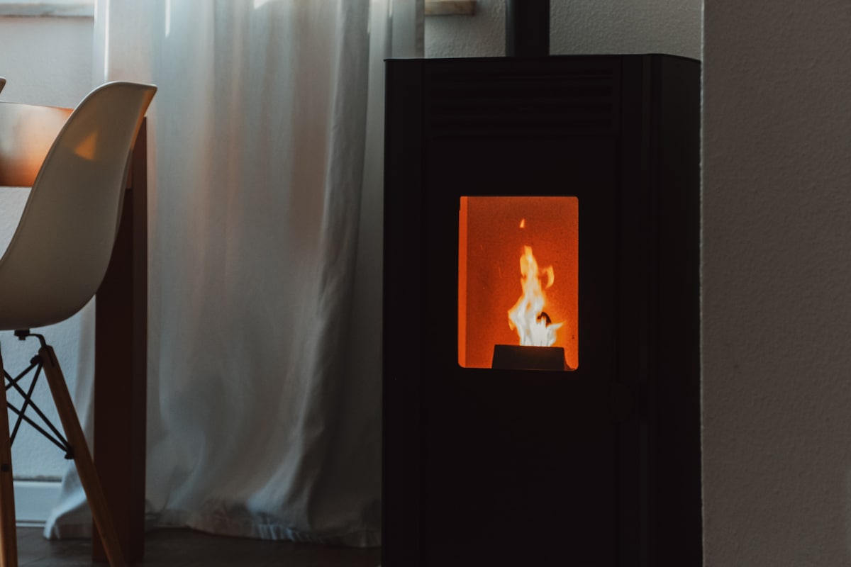 A modern stove is positioned near a wooden table, casting a warm glow through its glass panel. Soft, natural light filters in from a nearby window, with light fabric curtains gently framing the scene. The overall ambiance suggests a cozy indoor setting.