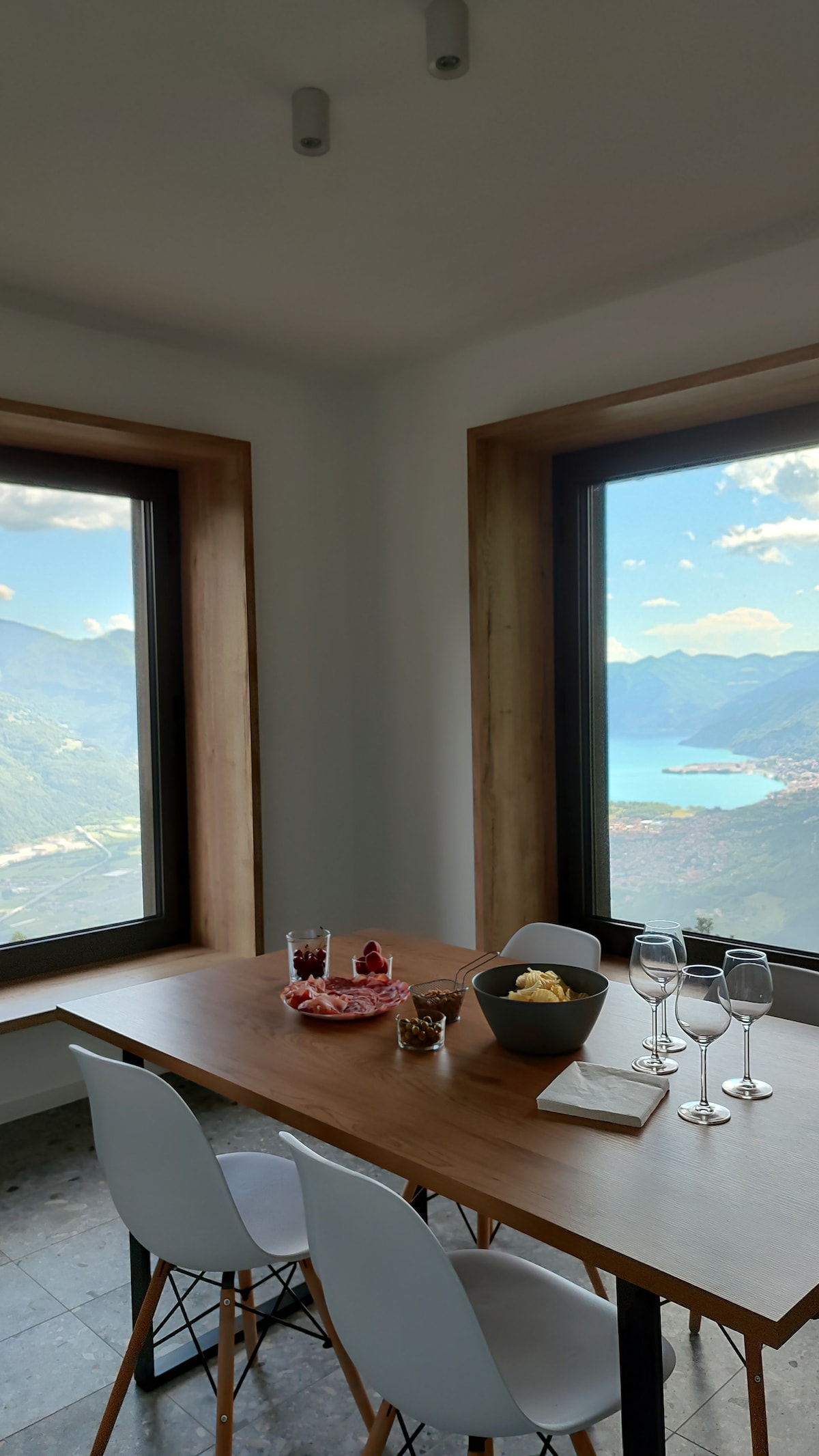 A dining area features a wooden table surrounded by white chairs. Two large windows frame scenic views of the lake and mountains. The table is set with a bowl of snacks, glasses, and a small plate. Soft natural light illuminates the space.