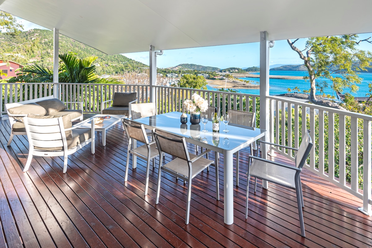 A spacious outdoor deck features a large dining table surrounded by modern chairs. A comfortable seating area is positioned nearby. Views of the Coral Sea and surrounding landscape are visible, framed by lush greenery and distant hills.