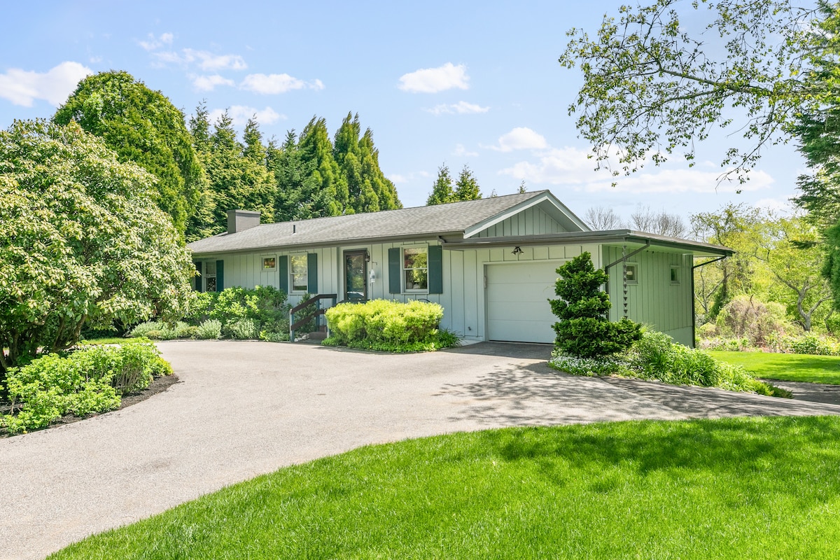 A single-story home is set within a lush green landscape, surrounded by trees and shrubs. A driveway leads to the front entrance, and a well-maintained lawn accentuates the peaceful atmosphere of the property.