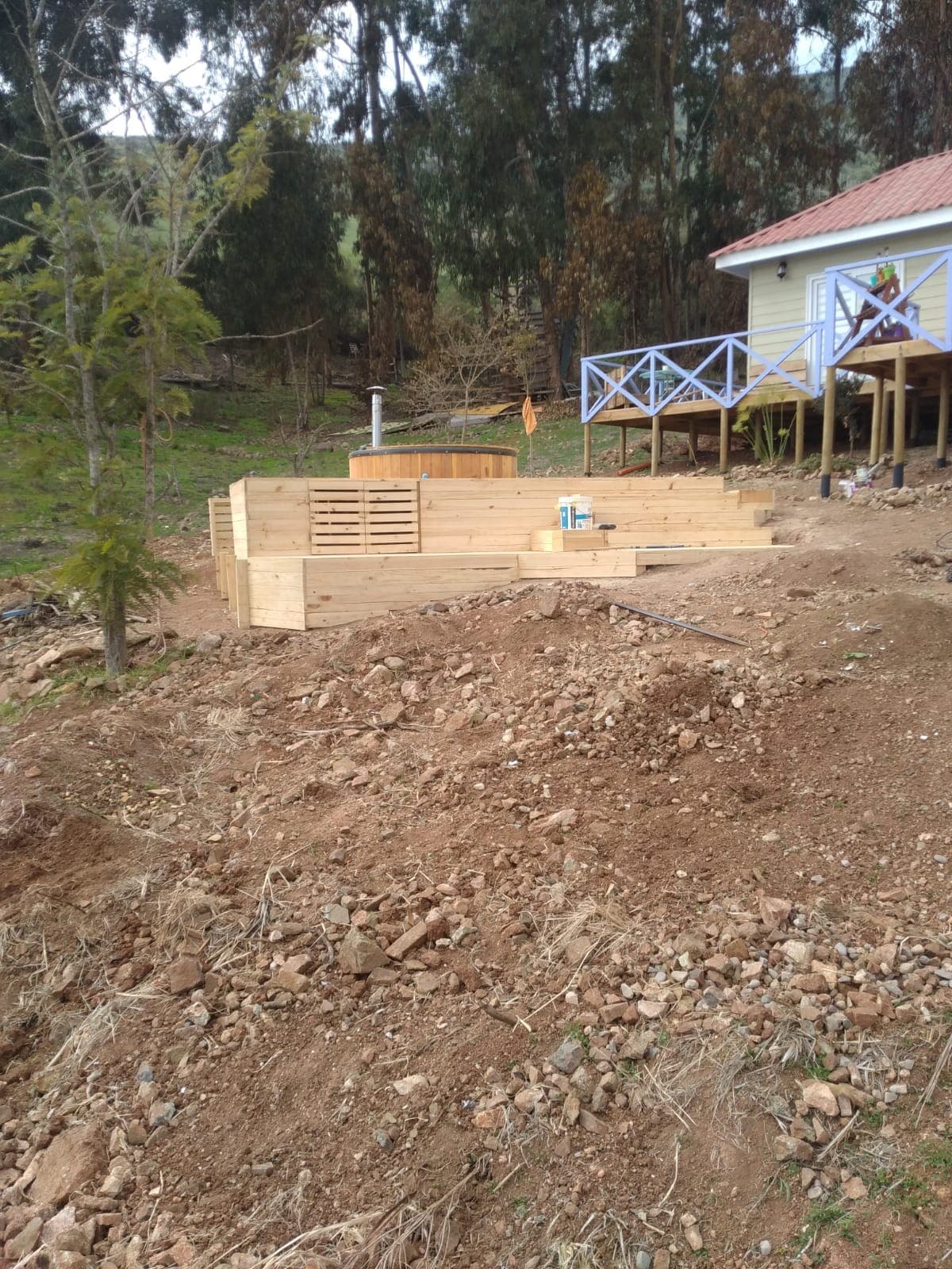 A garden area includes wooden platforms constructed from light-colored timber, designed for outdoor relaxation. Surrounding greenery features trees that provide shade, while a small building with a sloped roof stands in the background. A rough gravel surface is visible in the foreground.