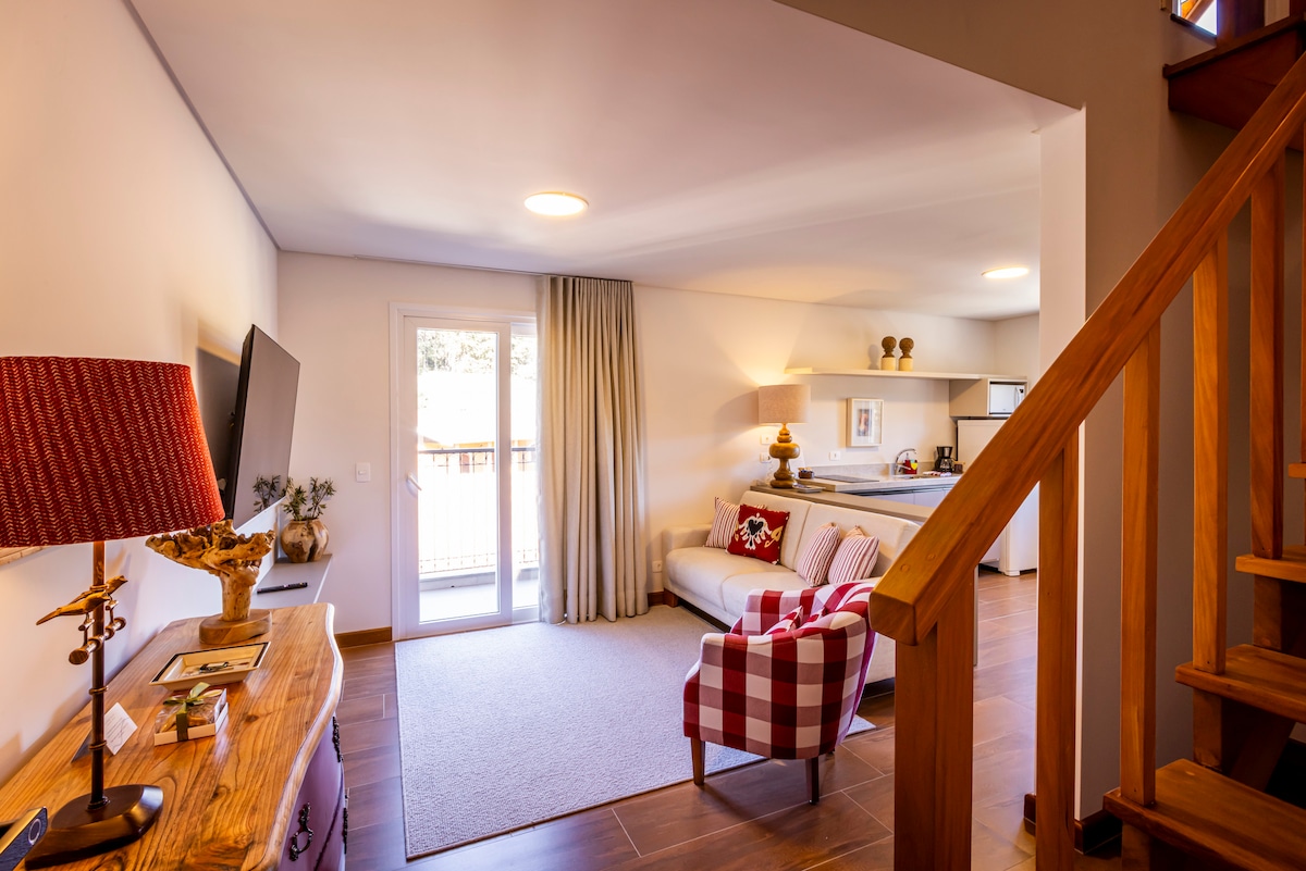 A spacious living area features a comfortable sofa and a decorative chair. Natural light flows through the sliding glass doors that lead to a balcony. A wooden console with a lamp and a large TV are positioned against the wall, enhancing the cozy atmosphere.