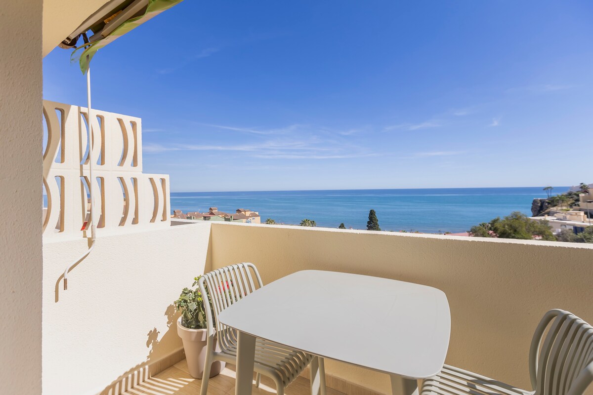 A private terrace is shown, featuring a light-colored table and chairs. The expansive view reveals the sea, with a clear blue sky above. A potted plant adds a touch of greenery, enhancing the outdoor space.