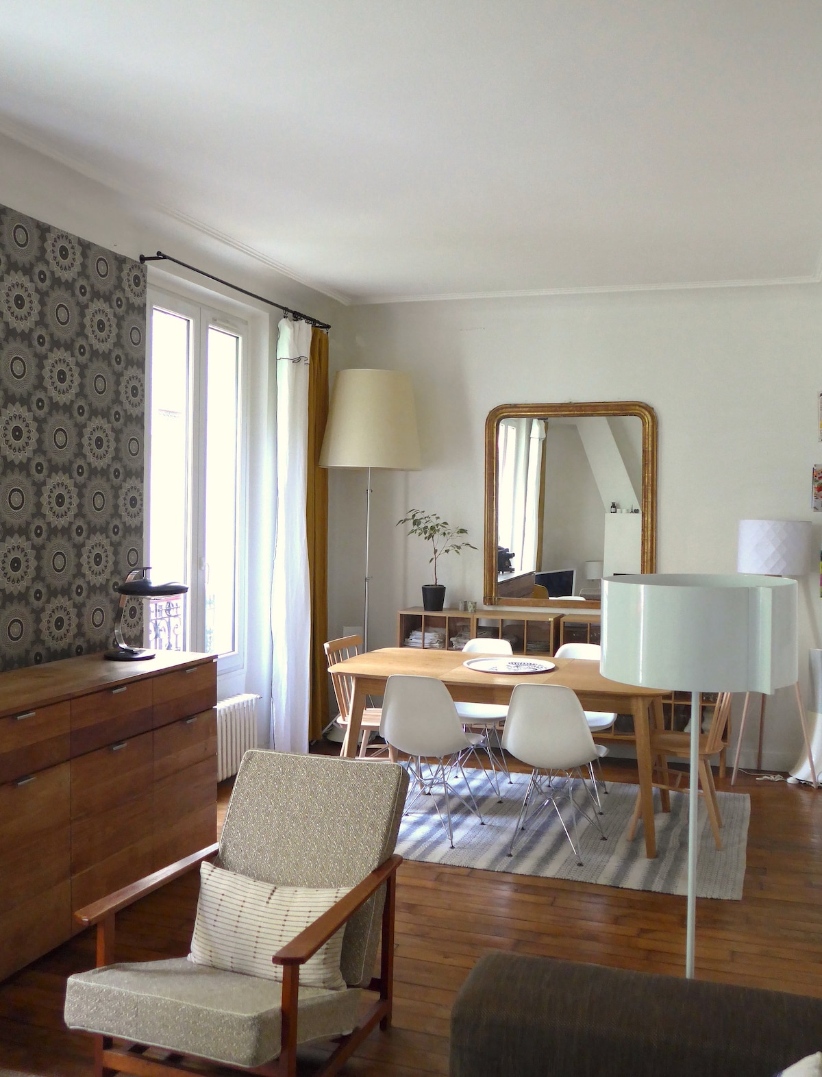 A light-filled living and dining area features a wooden dining table surrounded by white chairs. A vintage mirror reflects the space, with a tall floor lamp and potted plant adding character. Patterned wallpaper complements the warm wooden flooring.