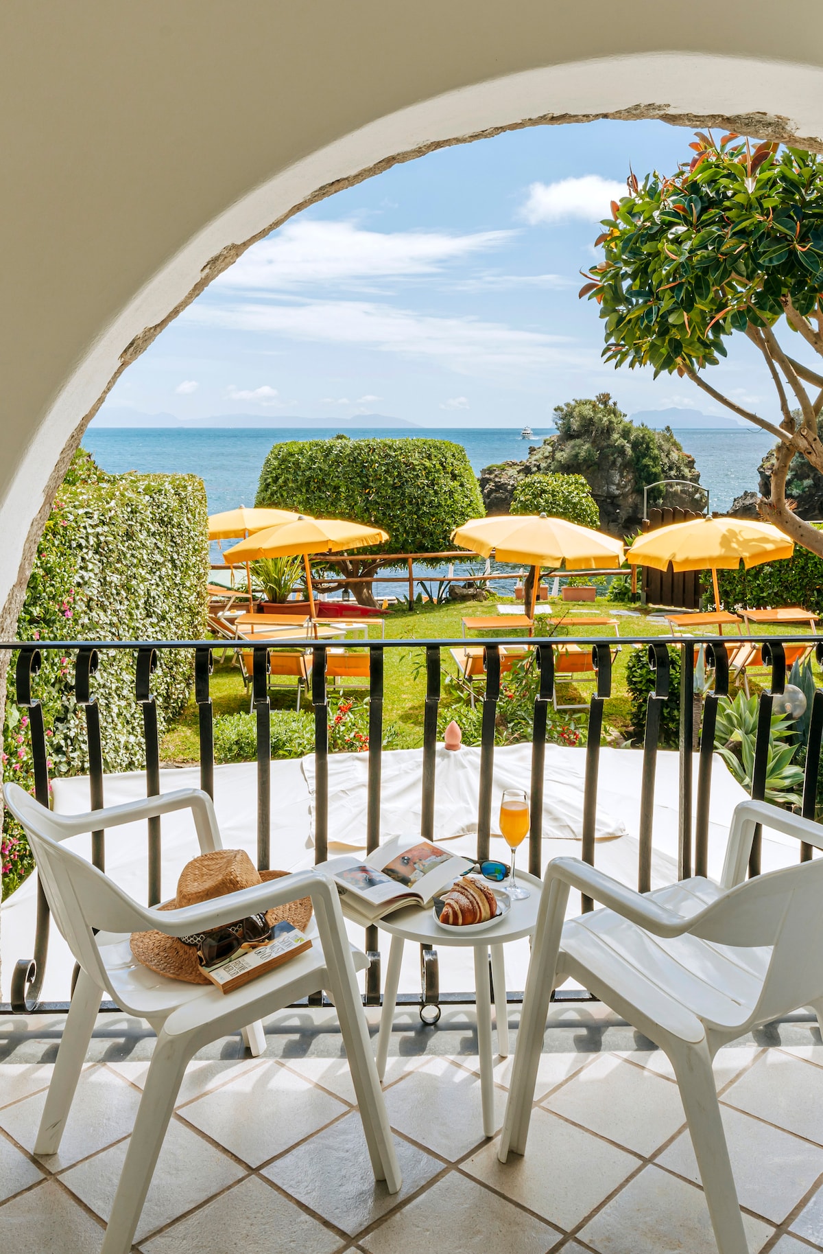 A view of a private terrace framed by an arch, overlooking a garden with yellow umbrellas and loungers. Two white plastic chairs are positioned with a straw hat and a book on a small table, creating a serene setting to enjoy the sea and garden scenery.