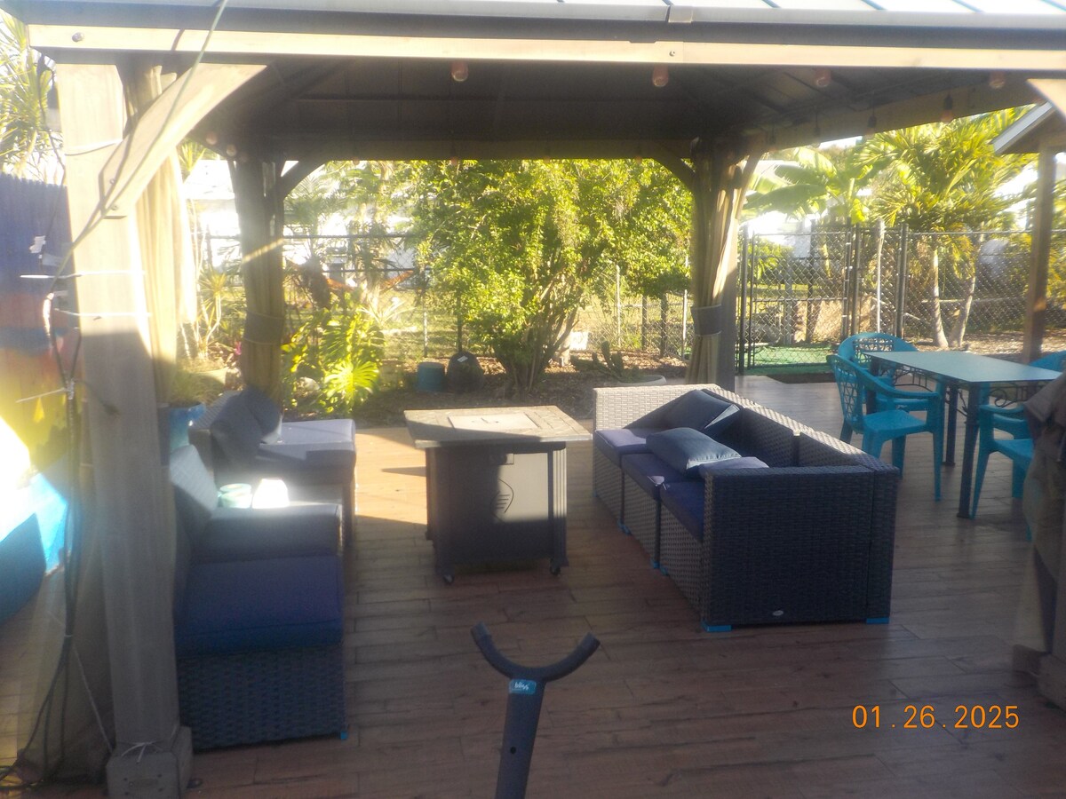 A covered outdoor seating area features a group of comfortable, modern sofas arranged around a coffee table. Surrounding greenery provides a natural backdrop, while a wooden deck enhances the inviting atmosphere of the space.