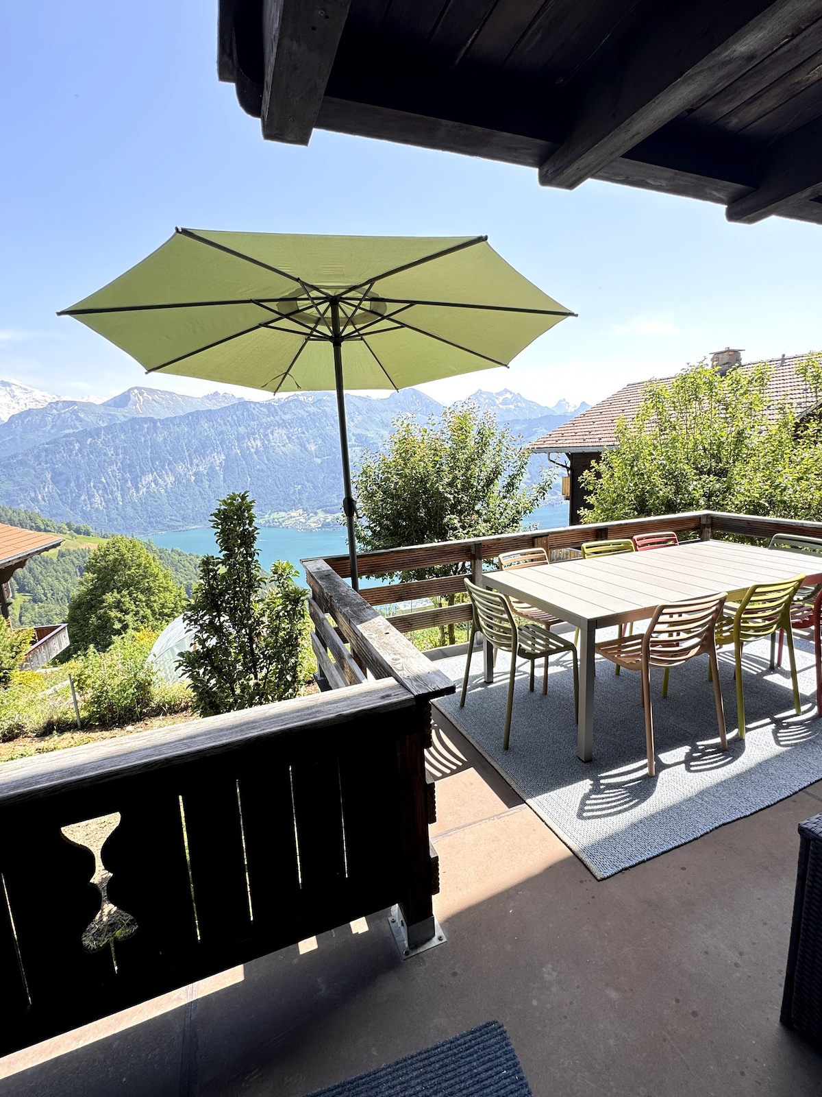 A spacious outdoor patio is presented, featuring a large table surrounded by colorful chairs. A green umbrella provides shade while offering views of the mountains and lake in the background. Lush greenery surrounds the area, contributing to a peaceful atmosphere.