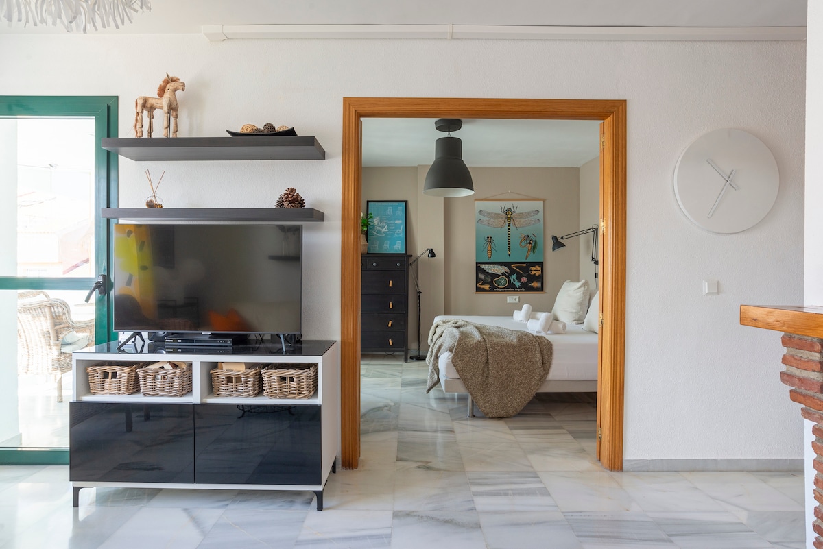 A modern living space featuring a sleek black and white entertainment unit with decorative baskets. A large television stands prominently. The room is connected to a cozy bedroom through an archway, illuminated by natural light from a nearby window.