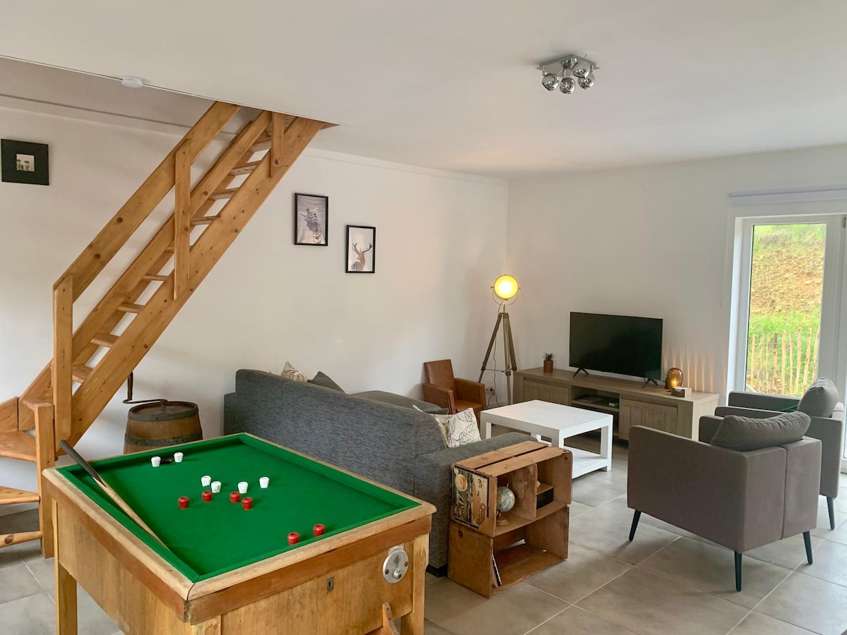 A welcoming living area is presented, featuring a green billiard table along one side. Two comfortable seating arrangements, including a gray sofa and armchair, are positioned around a coffee table, with natural light illuminating the space through a sliding glass door that opens to the outside.
