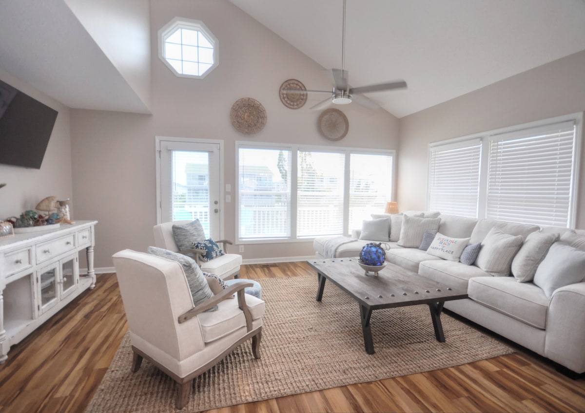 A spacious living area features large windows that allow natural light to fill the room. A light-colored sectional sofa and an upholstered armchair provide comfortable seating around a dark wooden coffee table. Decorative wall hangings add visual interest to the neutral-toned walls.