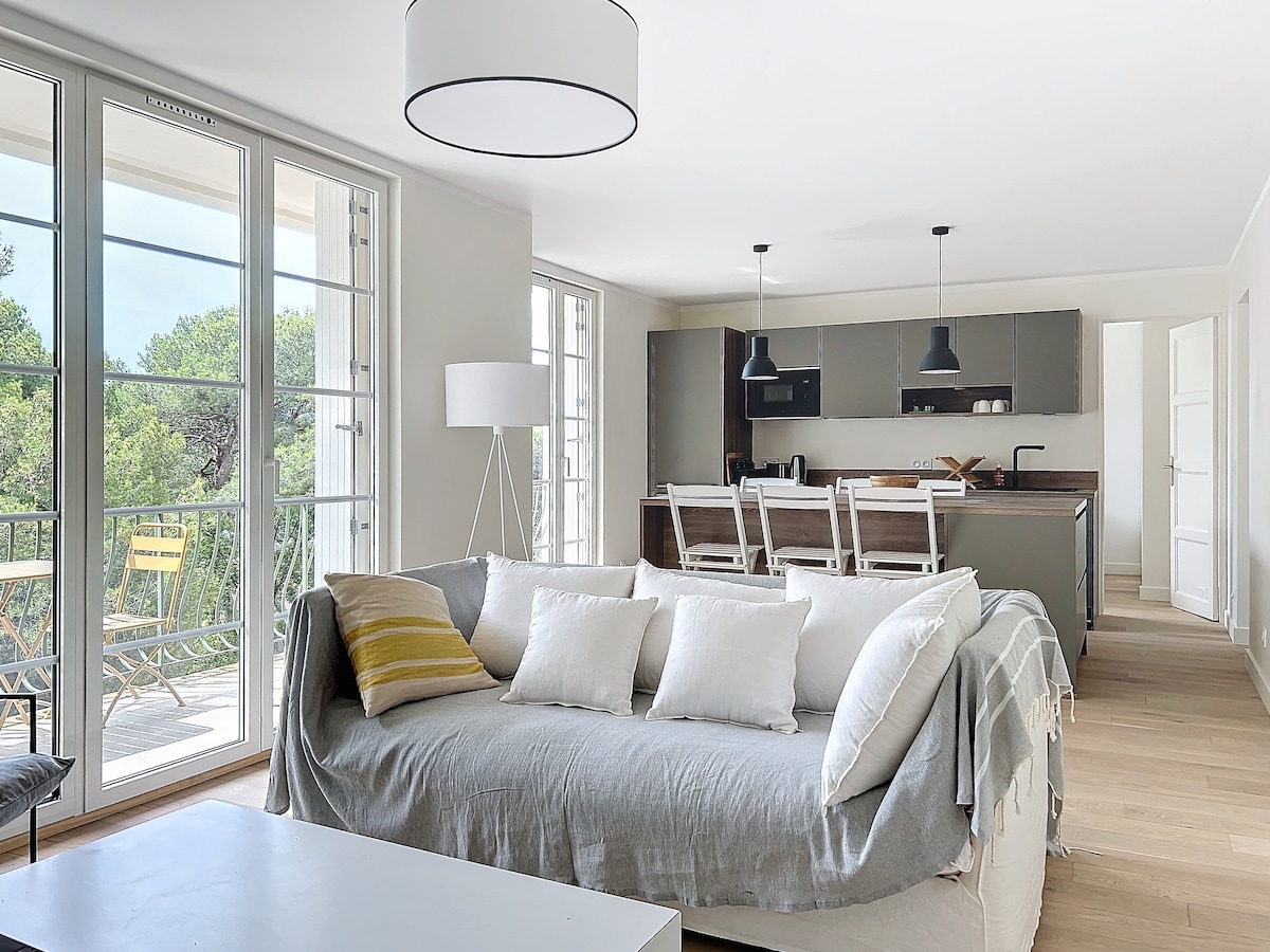 A spacious living area features a soft sofa adorned with multiple pillows. Large double doors open to a view of outdoor seating on a balcony. The kitchen area showcases modern cabinetry and bar stools, enhancing the open layout filled with natural light.