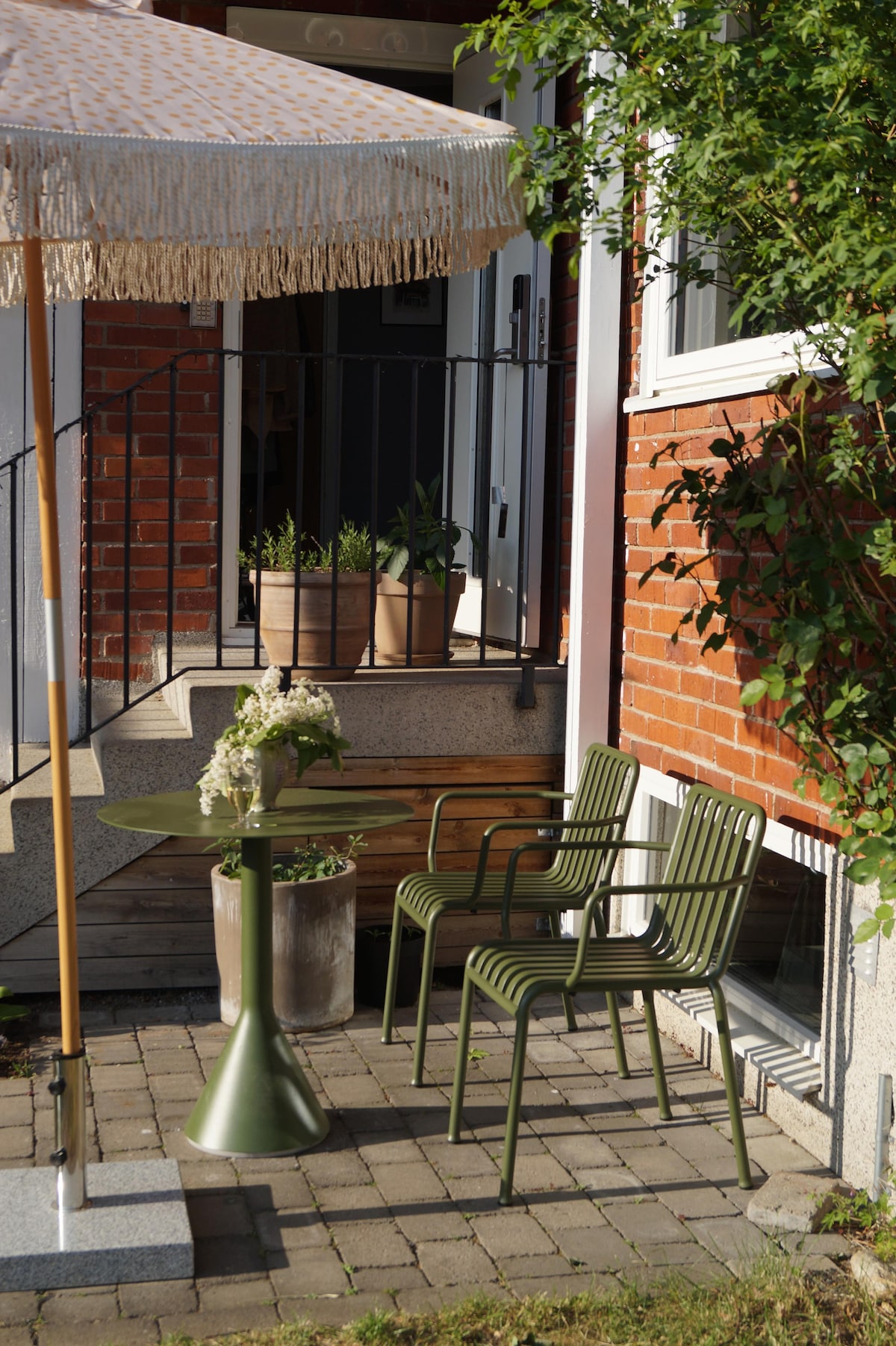 An outdoor seating area is set up with a round table and three green chairs, complemented by a parasol. Potted plants are positioned at the base of the steps leading to the entrance, with a brick facade adding texture to the surroundings.