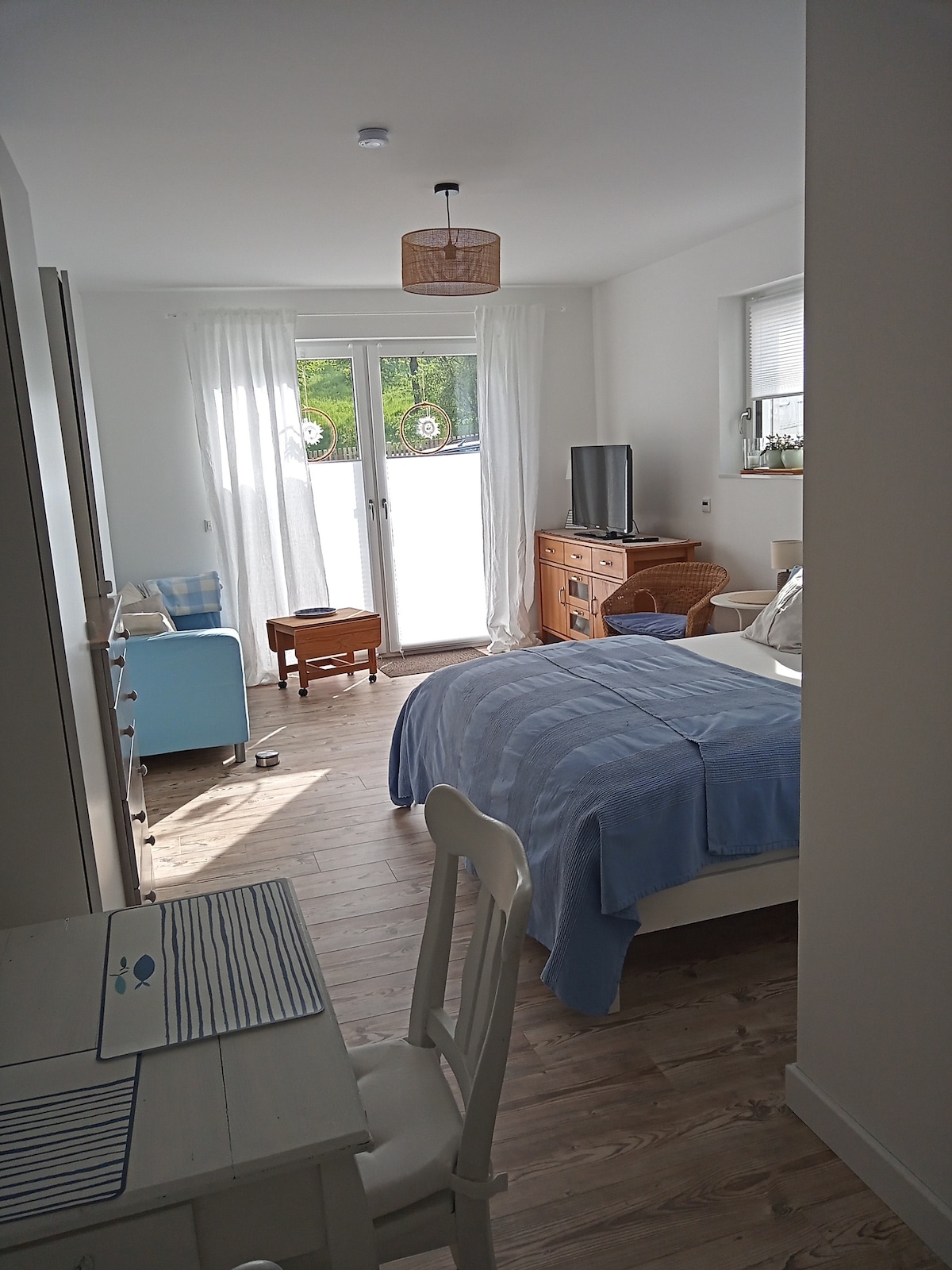 A well-lit studio room features a spacious bed covered with light blue bedding. Wooden flooring adds warmth, while large glass doors lead to an outdoor area. A small table and chairs, a television, and rustic furniture complete the inviting setup.