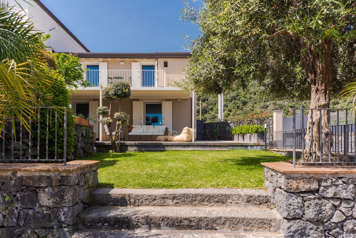 The exterior view of the two-story villetta features a well-maintained garden with greenery and stone steps leading up to the entrance. The covered terrace is visible, furnished with seating, providing an inviting space for relaxation. The surrounding area showcases a serene hillside backdrop.