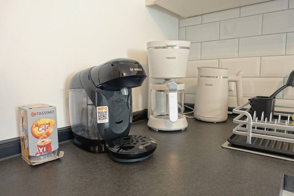 A small kitchen countertop features a modern coffee maker alongside a sleek kettle and a traditional coffee maker. A box of coffee pods is positioned nearby, and an organized dish rack is visible, providing a functional space for preparing beverages.