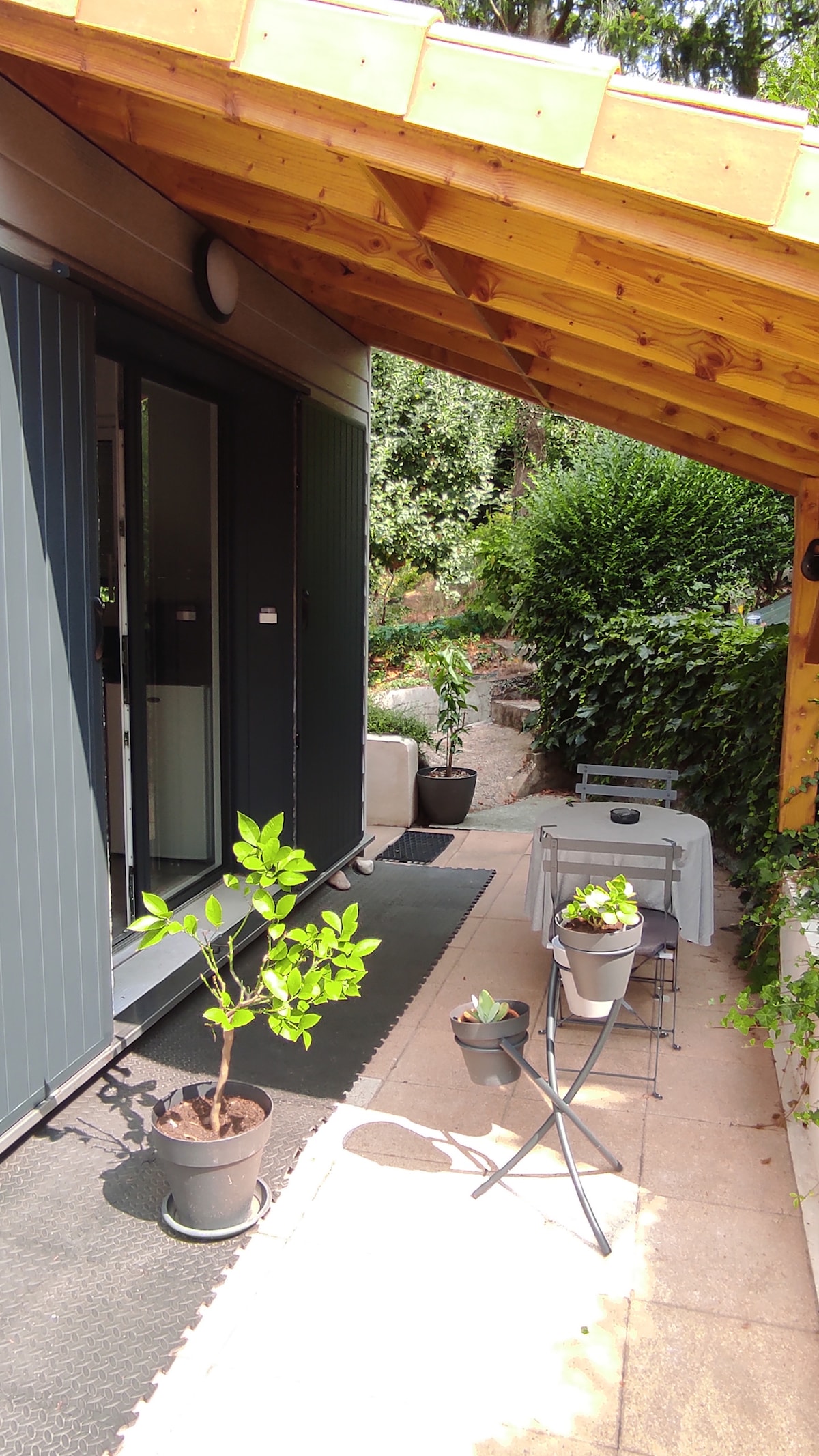 An entryway is highlighted, featuring a covered porch and potted plants. A small folding table with two chairs is positioned beside the entrance. Lush greenery can be seen in the background, contributing to a serene outdoor environment.