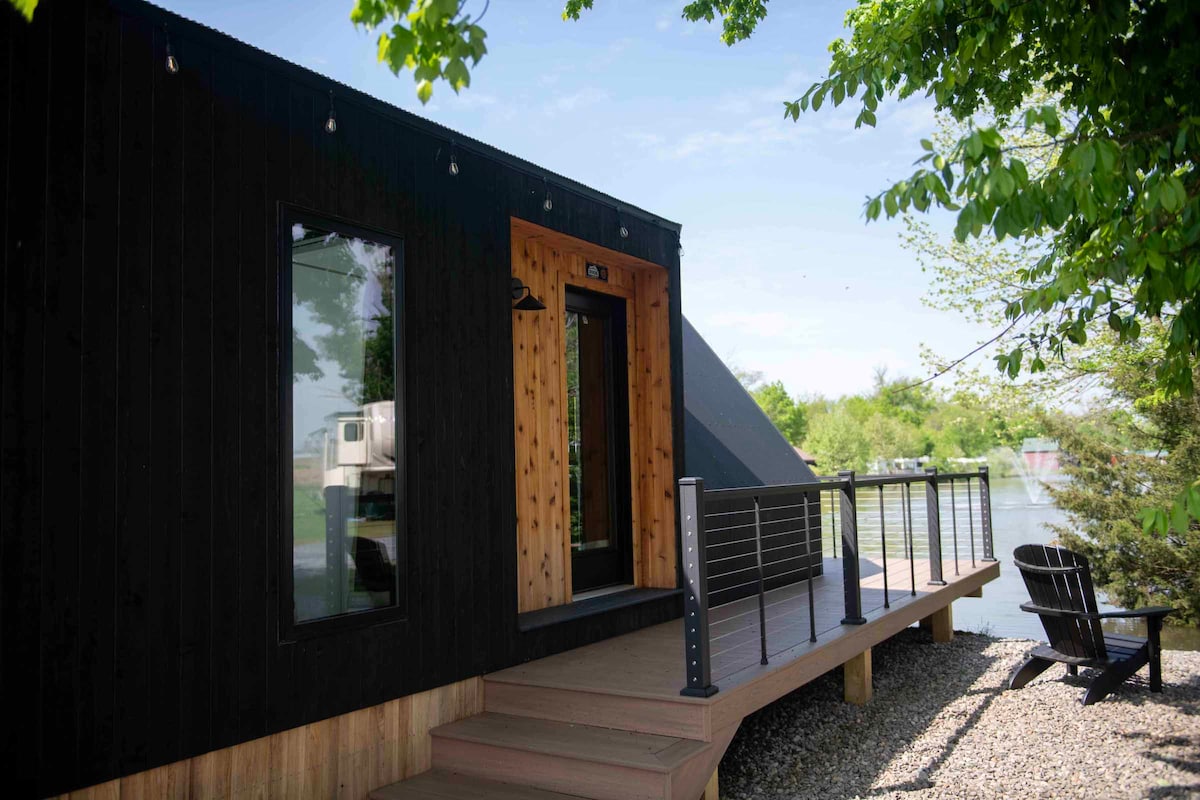 A modern A-Frame Tiny Home is set against a serene lakefront backdrop. Large windows invite natural light, and a welcoming porch extends out, featuring a railing. Lush greenery surrounds the area, enhancing the tranquil outdoor environment.